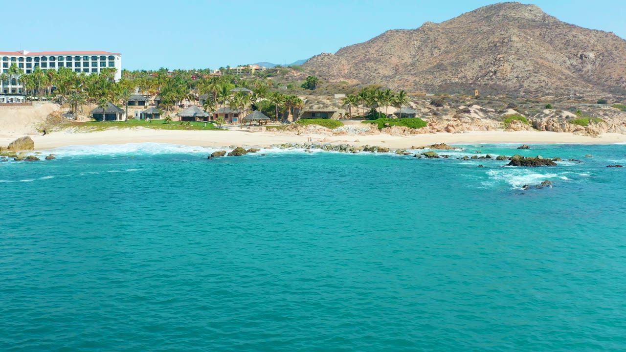 Clear ocean foreground with waves breaking near rocks below a beach and resort buildings.