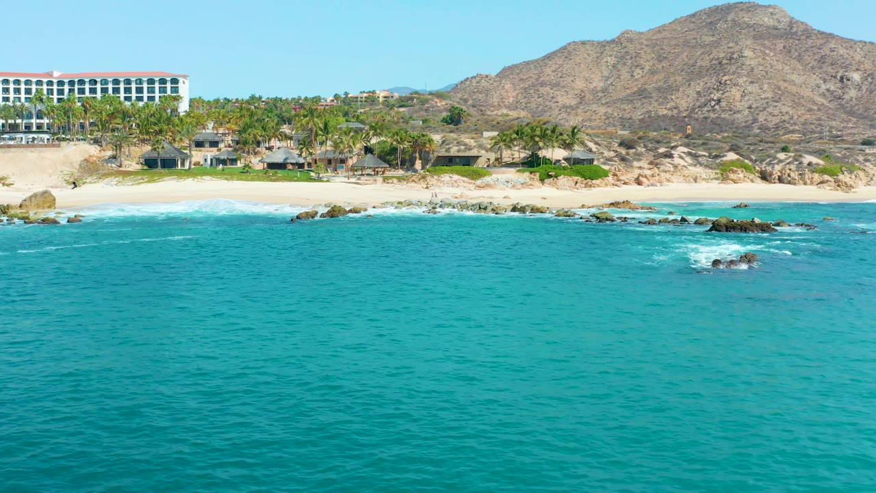 Elevated coastal frame from Marquis Los Cabos, Mexico showing foam spreading around rocks near the sandy shore, turquoise water across the foreground, palm-lined buildings, a white resort block, mountain ridge, and no visible people.