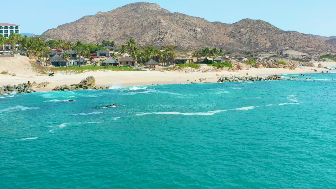 Wide aerial view at Marquis Los Cabos, Mexico with turquoise water in the foreground, a sandy beach with rocks and palms in the middle distance, buildings near the shore, mountains behind, and no visible people.
