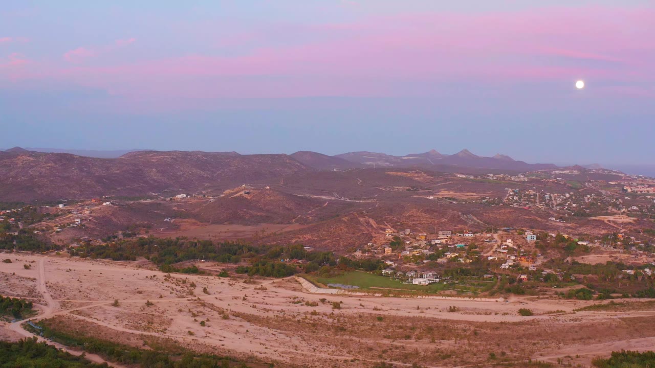 San Jose del Cabo landscape with pale dirt tracks in the foreground, clustered hillside buildings in the midground, distant ridges, a pink-blue sky, and no visible people.