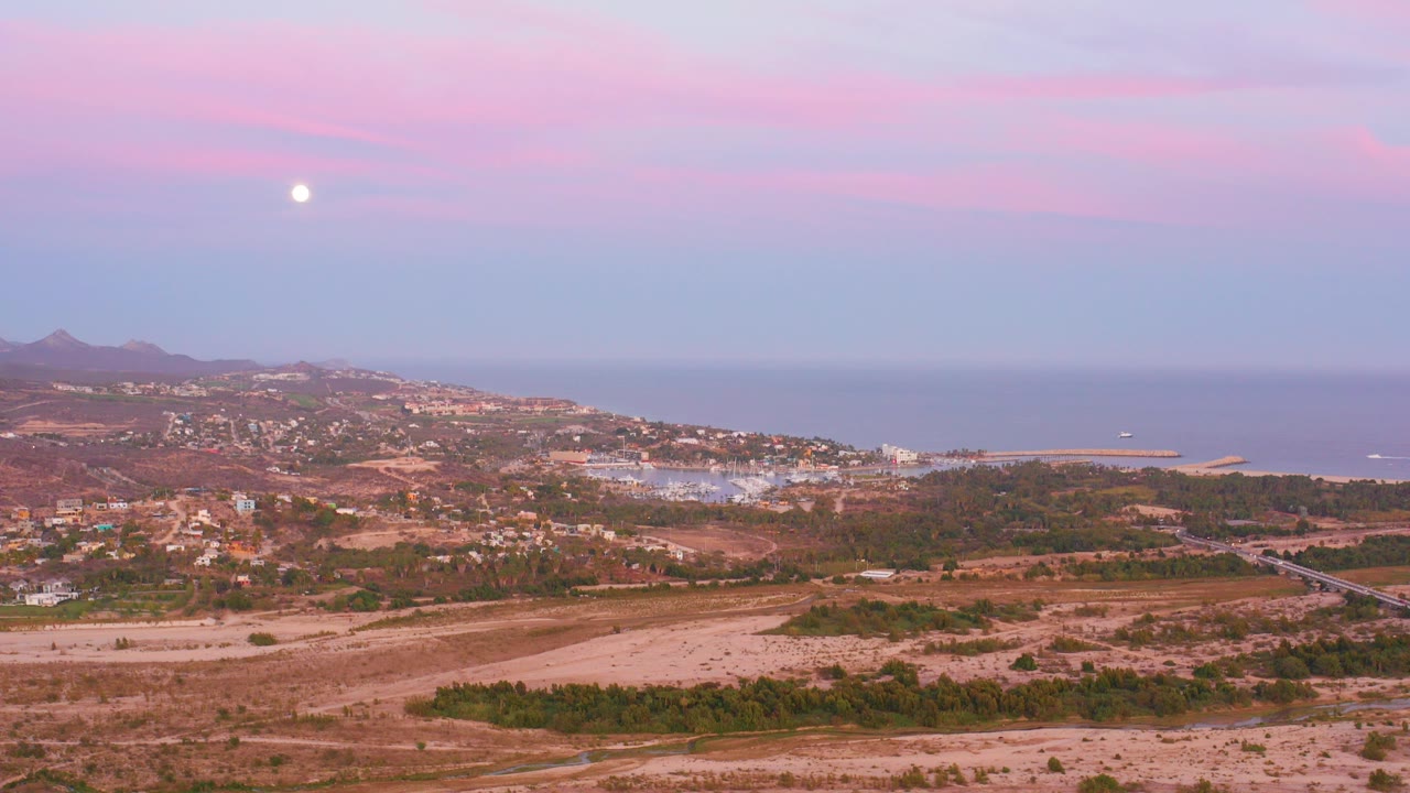 Wide San Jose del Cabo coastal landscape with dry fields, hillside buildings, bay water, distant mountains, a pink sky, the moon, and no visible people.