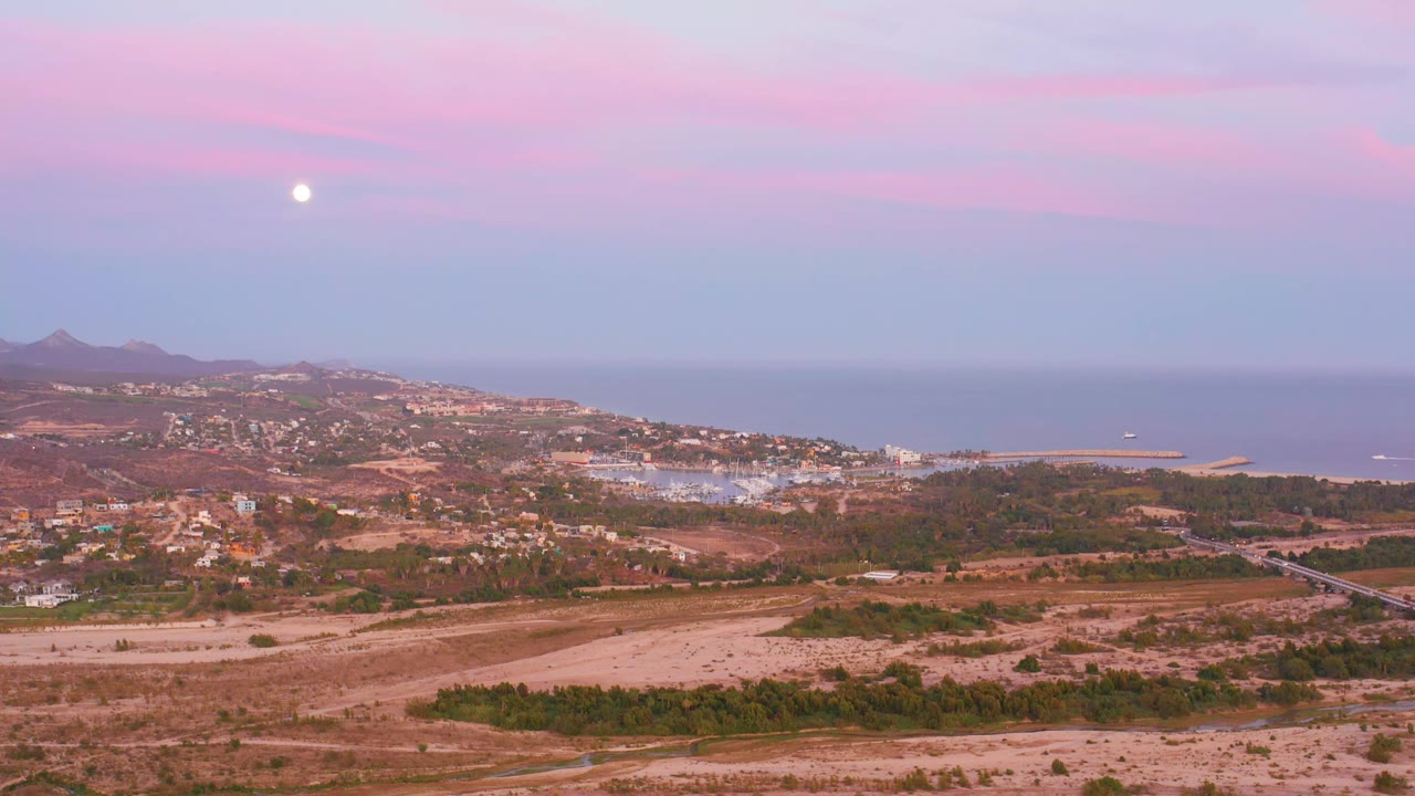 Coastal town and dry hills below a pale moon and pink clouds.