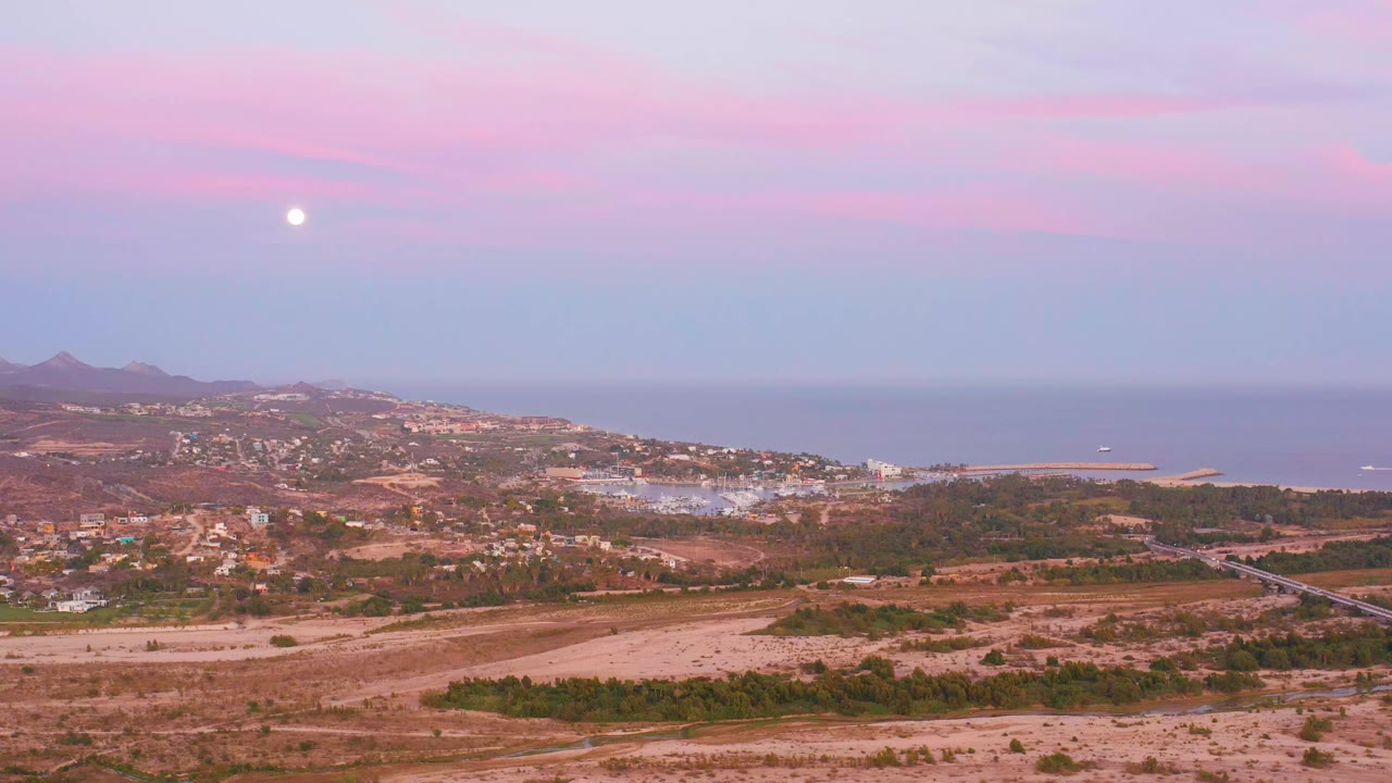 San Jose del Cabo coastal hills and scattered buildings sit below a pink-purple twilight sky with the moon above the ocean horizon, no visible people.