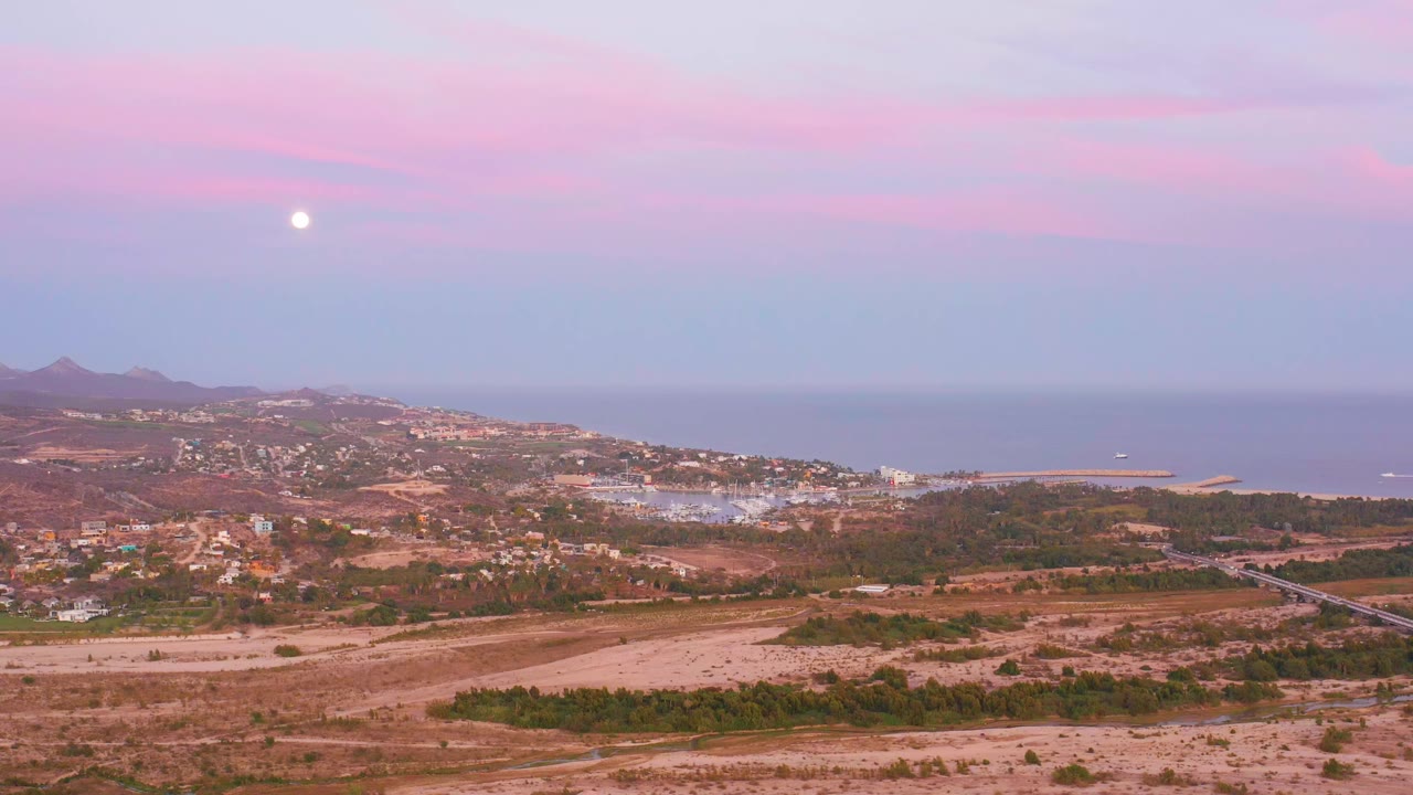 An elevated San Jose del Cabo view shows hillside buildings, reddish dry terrain, a road, the ocean horizon, and a bright moon in the twilight sky, no visible people.