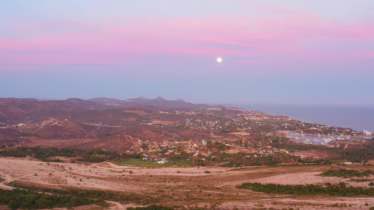 Opening frame of San Jose del Cabo hillside terrain with dry scrub, town buildings, distant ocean, low mountains, a pink sky, a bright moon, and no visible people.