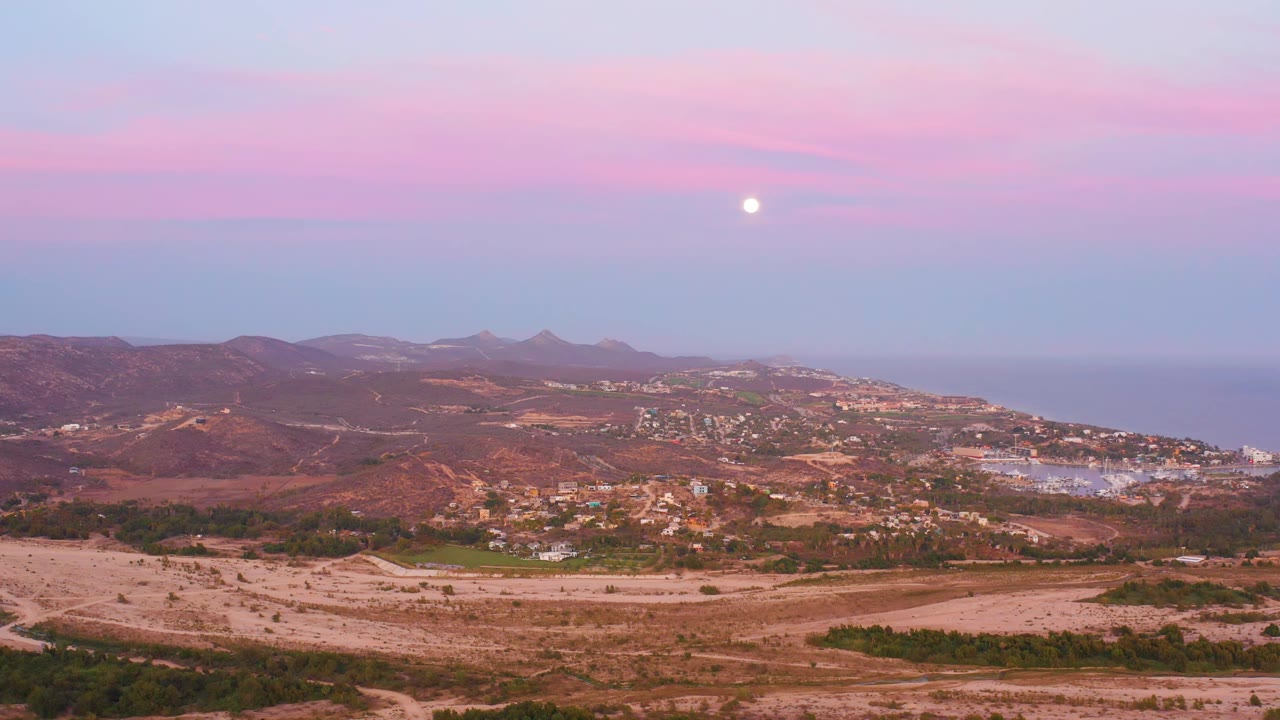 San Jose del Cabo hillside view with reddish dry hills, clustered buildings, a blue ocean horizon, pink cloud bands, the moon high in frame, and no visible people.