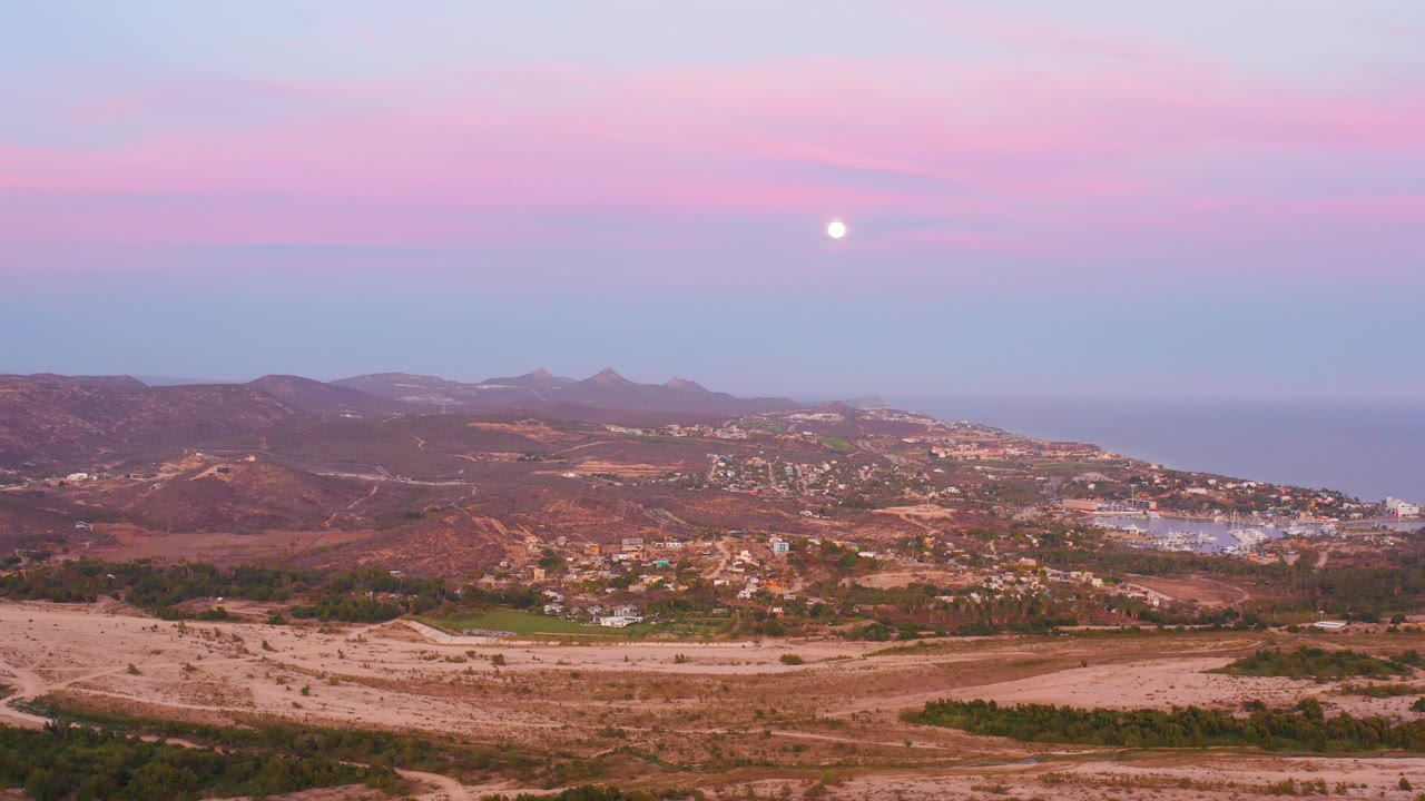Wide aerial-style San Jose del Cabo landscape showing pale foreground land, reddish hills, town structures, distant sea, low mountains, pink sky, moon, and no visible people.