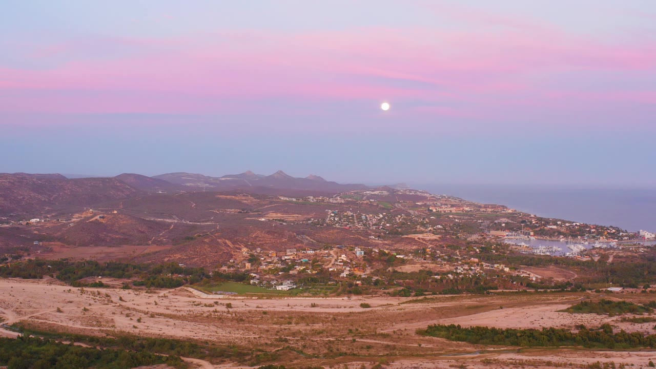 Pink sky and moon above hills, fields, town, and ocean