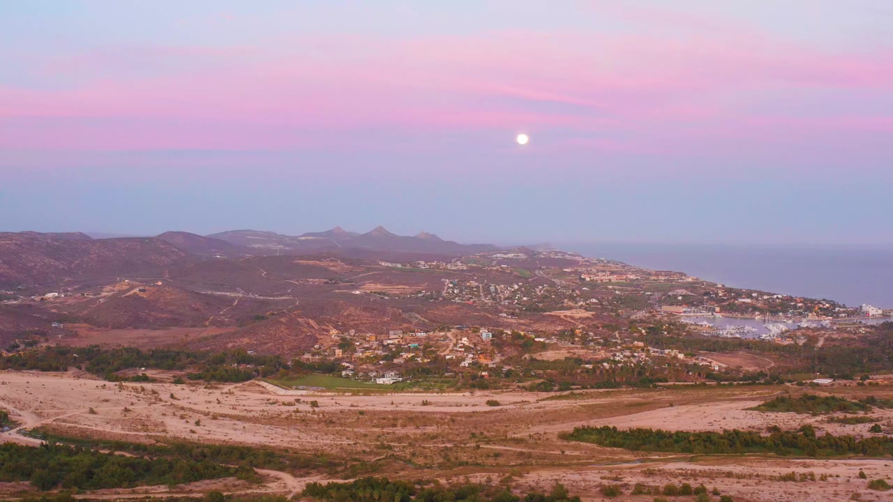 At 0:00, San Jose del Cabo appears from an elevated wide view with dry foreground terrain, hillside town, distant ocean, pink dusk sky, and no visible people.