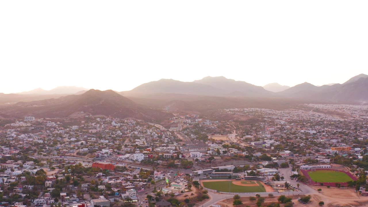 San Jose del Cabo aerial frame with the baseball diamond and green field in the lower right, compact urban buildings across the middle, dry hills behind them, bright sunset haze, and no visible people.