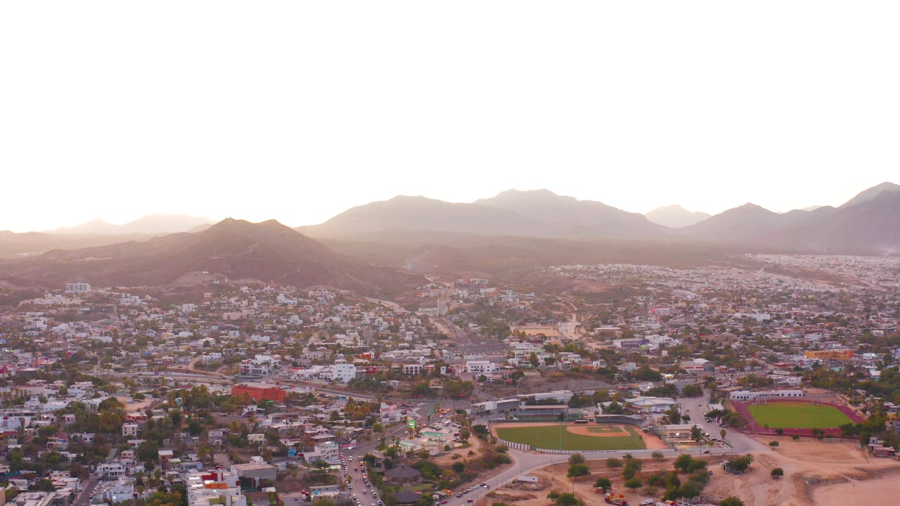 San Jose del Cabo aerial view at the start of the clip, showing dense low-rise neighborhoods, roads, green sports fields, hazy mountains, warm dusk light, and no visible people.