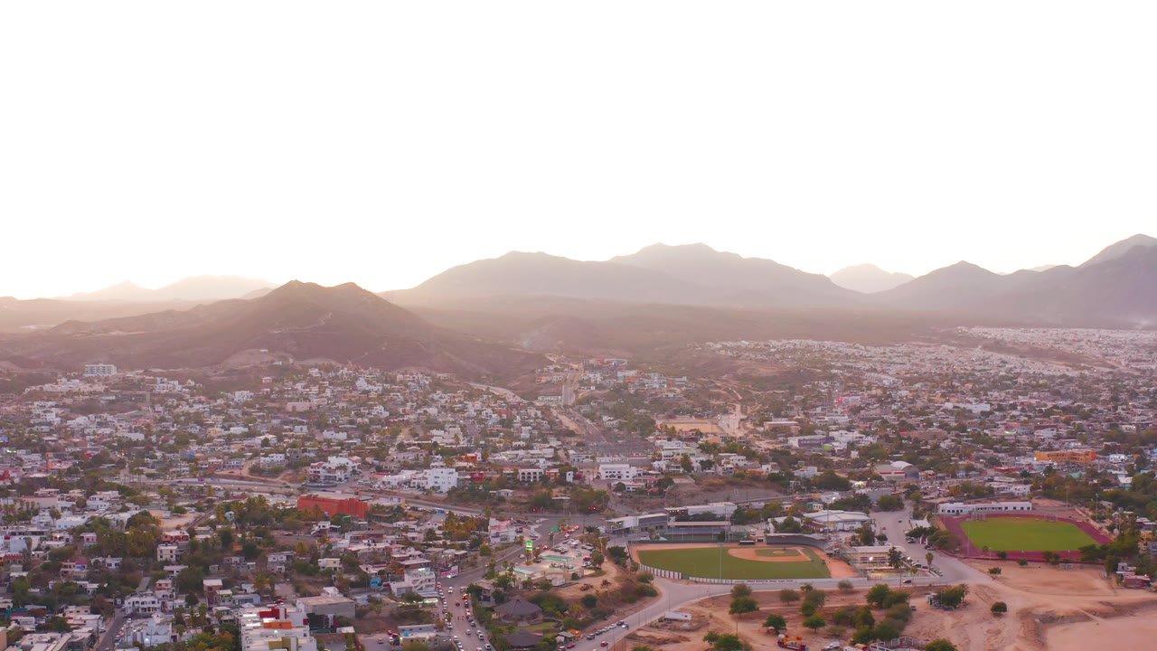 Wide aerial view over residential blocks and sports fields with mountains under a bright hazy sky.