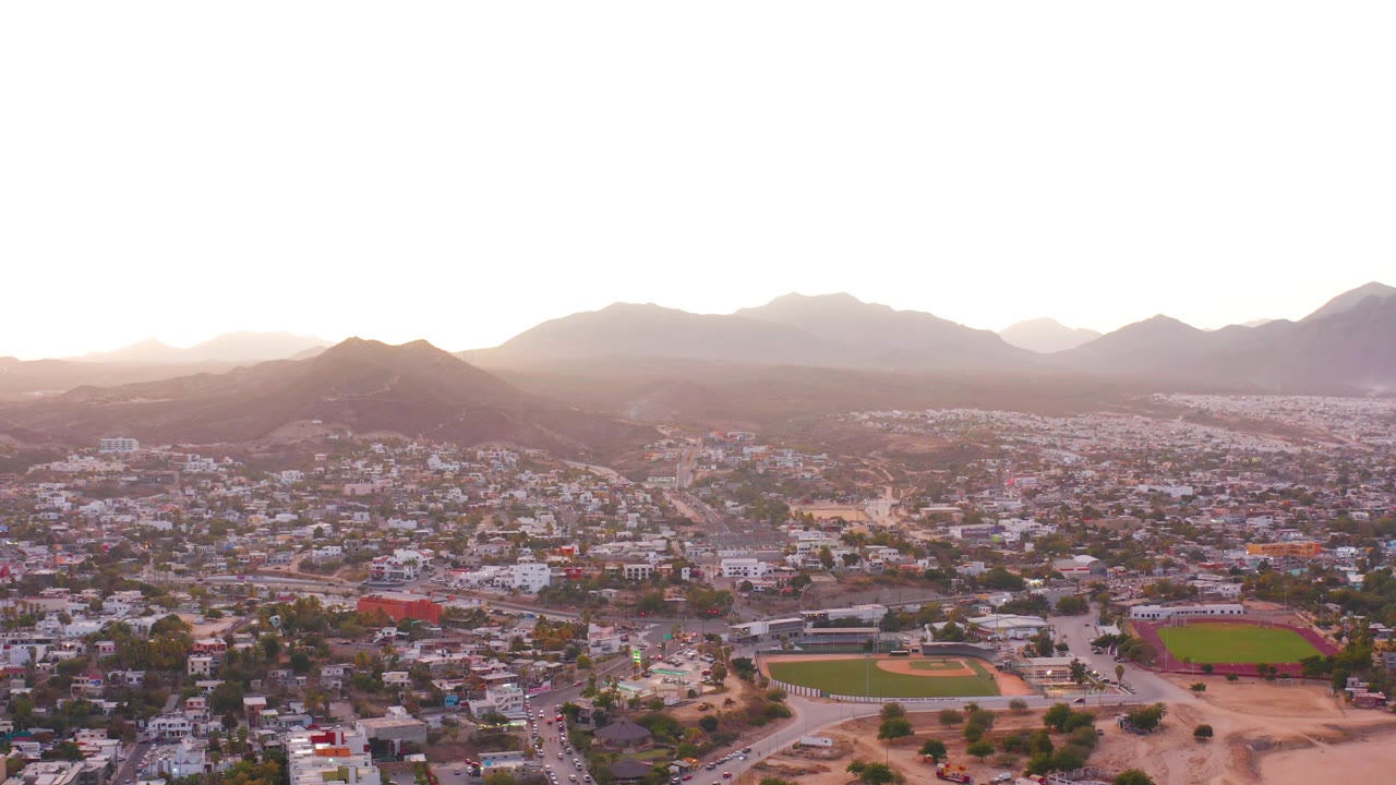 San Jose del Cabo is seen in a slightly later dusk frame with dense buildings below, green sports fields to the right, layered mountains behind the town, and no visible people.