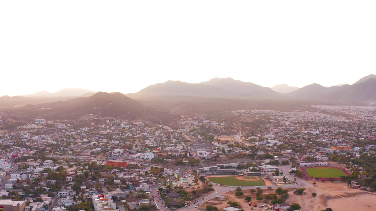 Wide aerial view over San Jose Del Cabo showing residential blocks, roads, two sports fields, dry open ground, hazy mountains, and no visible people at the start of the clip.