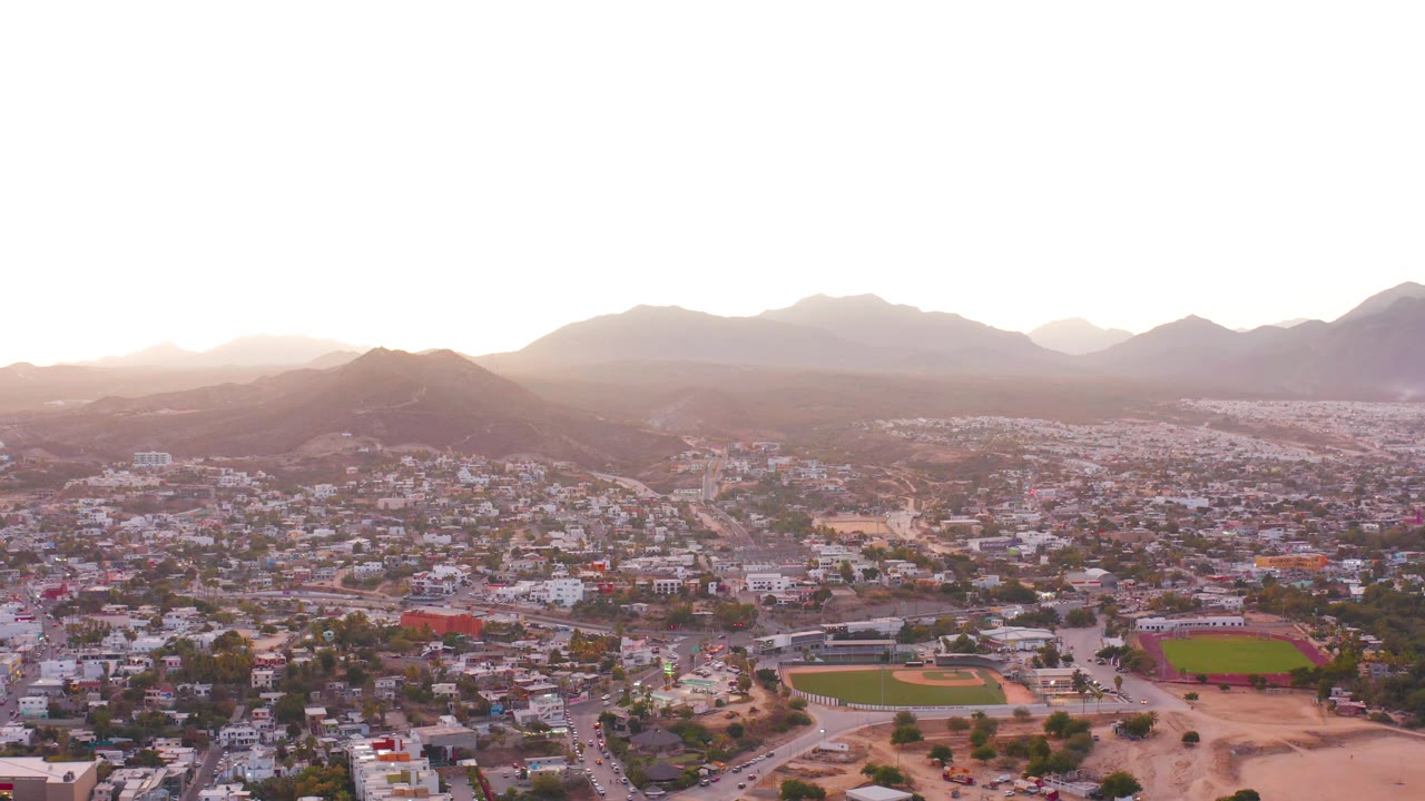 Aerial frame over San Jose Del Cabo with the baseball diamond and green field near the lower center, dense low-rise buildings beyond, mountains on the horizon, and no visible people.