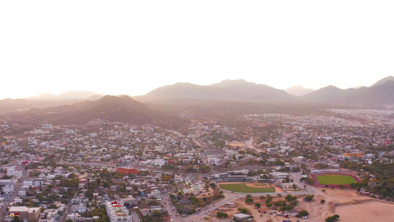 Wide drone view over low-rise buildings, fields, and mountains.