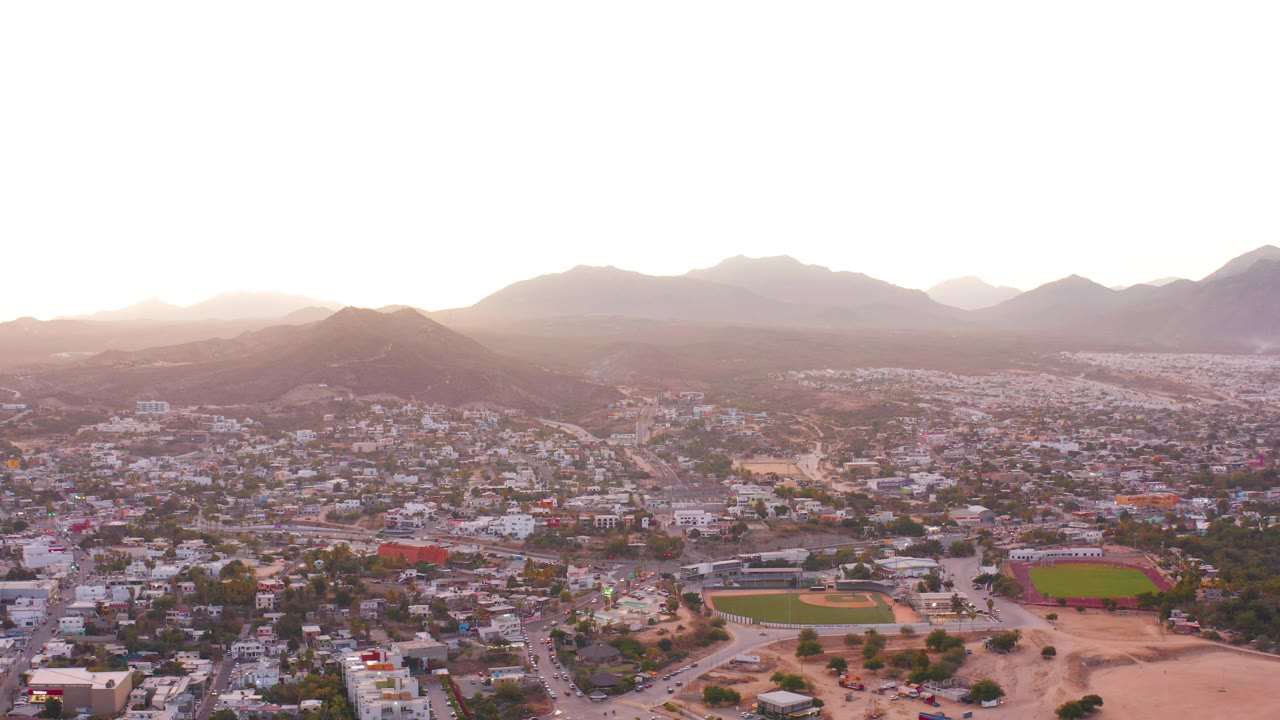 Aerial frame over San Jose del Cabo with white and tan buildings, a green rectangular field, a baseball field, dusty foreground lots, and backlit mountains under a pale sky with no visible people.