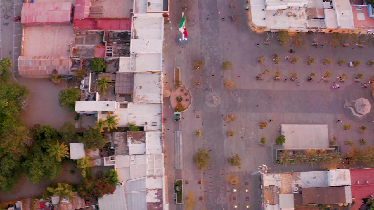 Top-down town square with flag, trees, rooftops, and street edges.