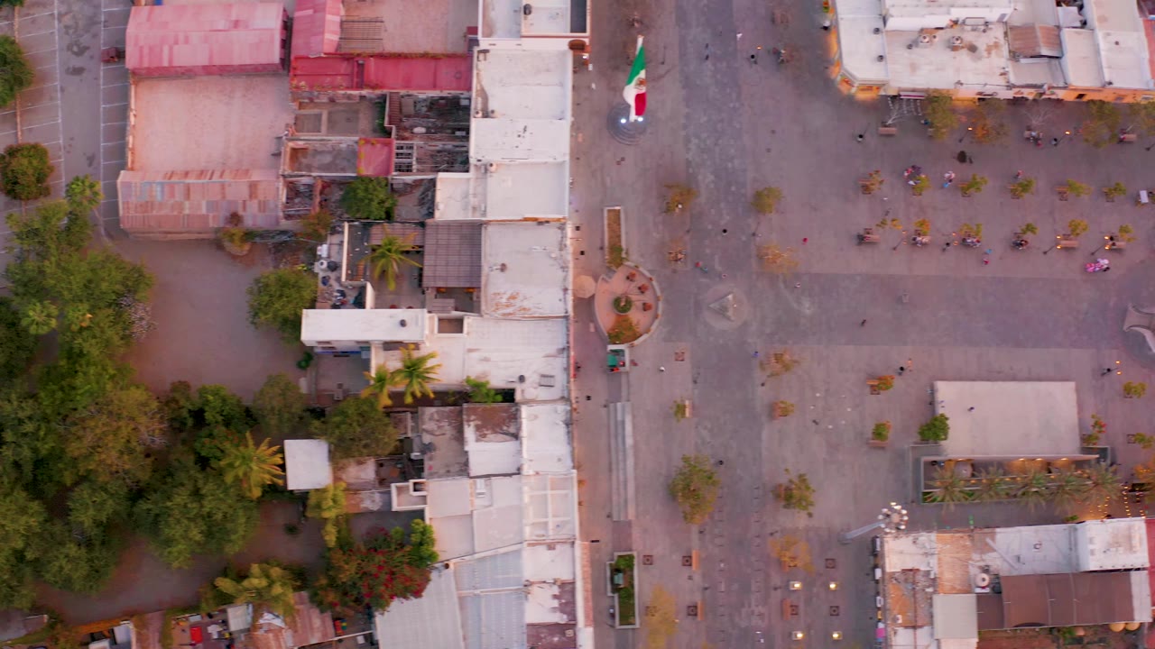 Aerial San Jose del Cabo view with the paved plaza across the right side, a Mexican flag near the upper road, buildings along the left edge, green trees below, and no visible people.
