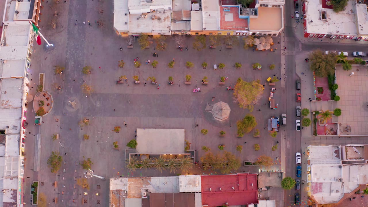 Aerial view of the plaza with the flagpole, central structure, trees, streets, and roofs.