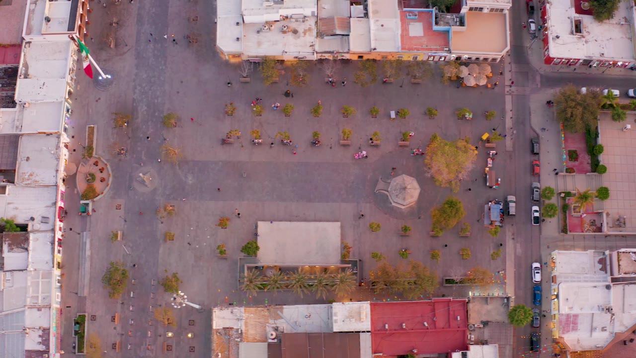 The rotating overhead view of the San Jose del Cabo town square places the flag near the upper left, with tree rows, a central structure, vehicles along the street, and no visible people.
