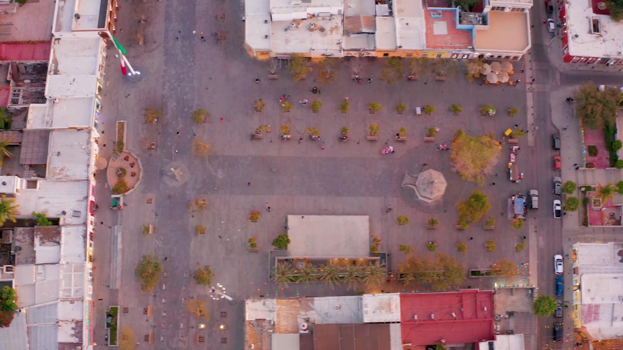 Rotated aerial frame of the square with flagpole, buildings, trees, and open pavement.