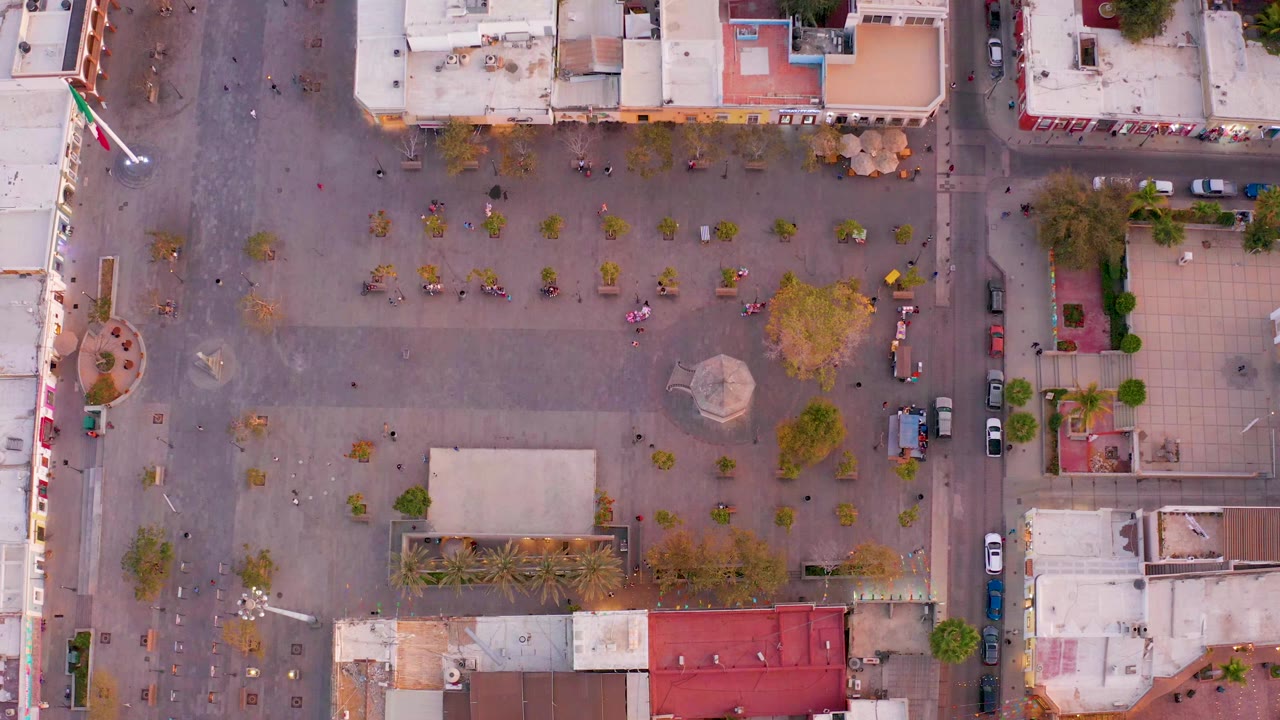 Overhead frame of a San Jose del Cabo town square at the start of the clip, showing scattered people across the paving, small trees, surrounding roofs, parked cars, and a flag along the left edge.