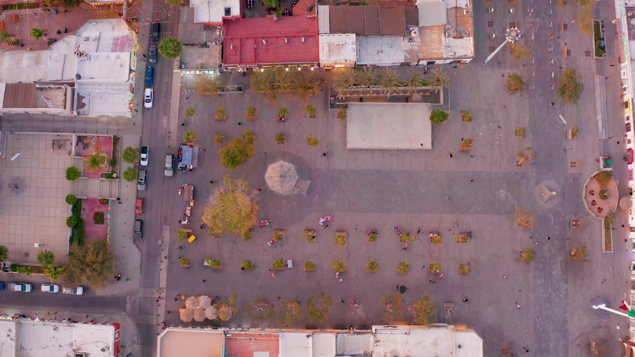 San Jose del Cabo plaza from above, showing the central paved open area, a red-roofed building, tree rows, curbside cars, and no discernible people.