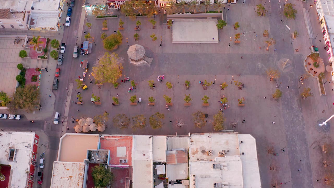 Aerial plaza view with a central circular structure, trees, cars, and surrounding rooftops.