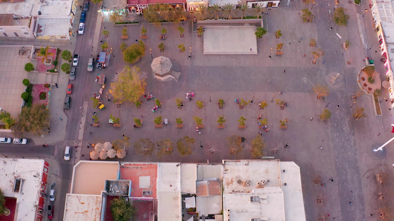 Drone-style overhead frame of San Jose del Cabo showing the plaza, a light-colored rectangular paved area, scattered trees, bordering streets, rooftops, cars, and tiny people.