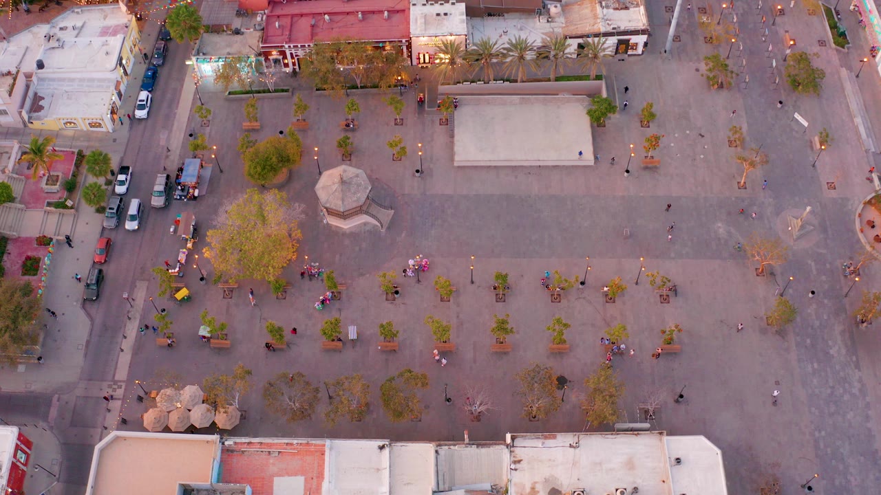 Overhead view of a San Jose del Cabo town square with scattered trees, a small central structure, parked cars along the side street, and pedestrians spread across the plaza.