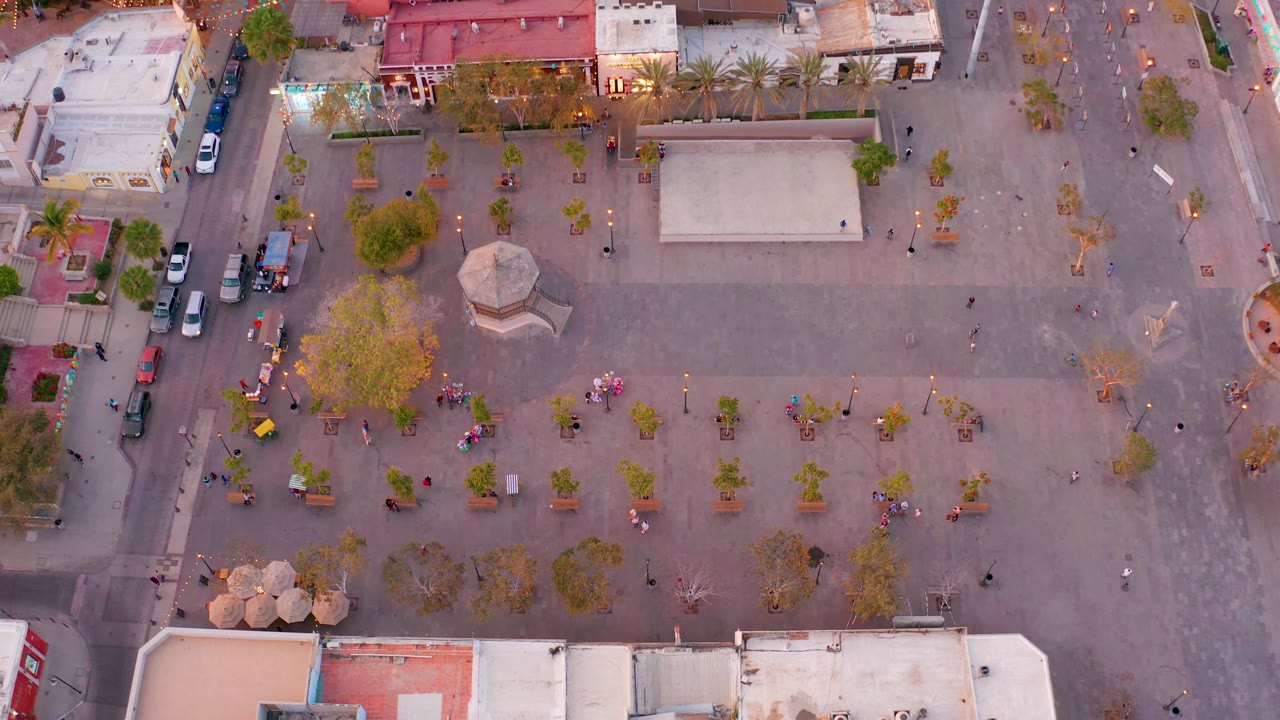 Aerial plaza view with pedestrians, trees, buildings, and cars near the curb.