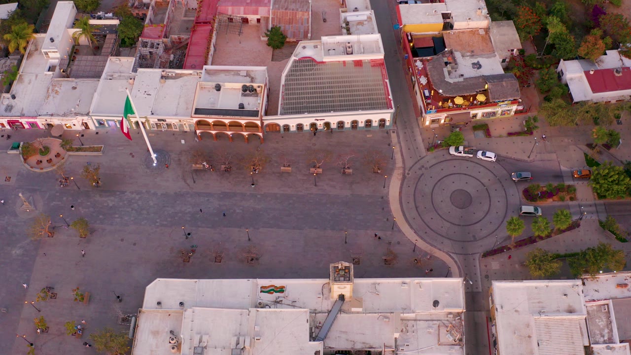 Overhead view of a San Jose del Cabo plaza at the start of the clip, with the Mexican flag extended left of center, a circular roundabout on the right, white rooftops, trees, cars, and tiny pedestrians.