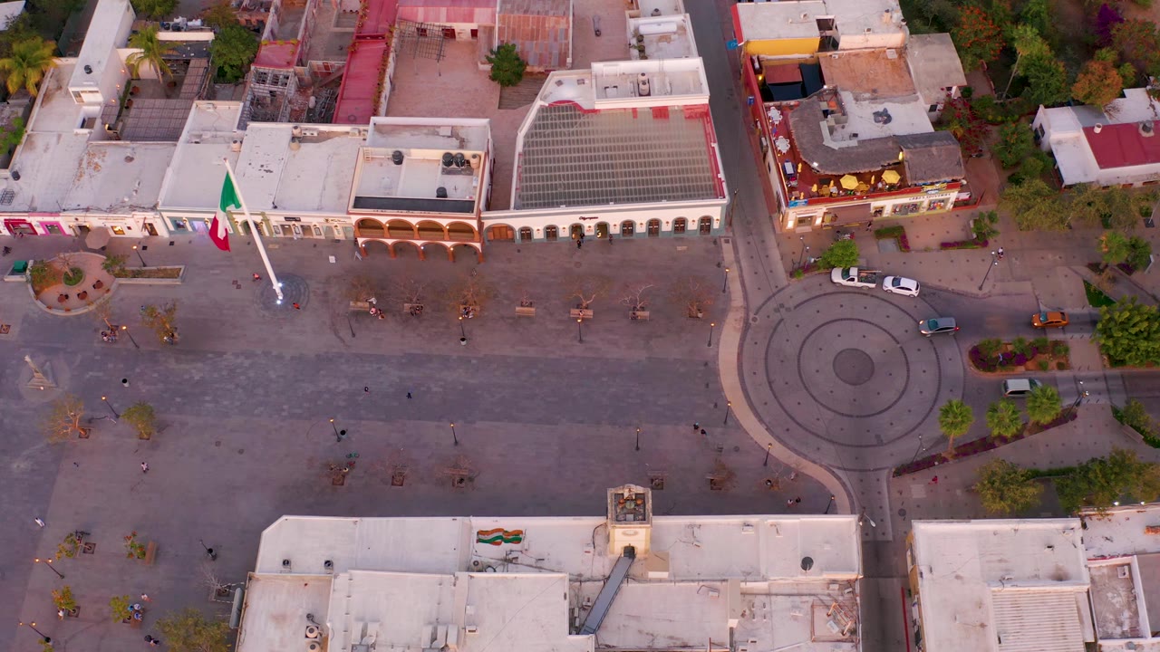 Overhead San Jose del Cabo plaza view with the Mexican flag near the left side, the circular road and central island on the right, low buildings around the square, a few vehicles, and scattered pedestrians.