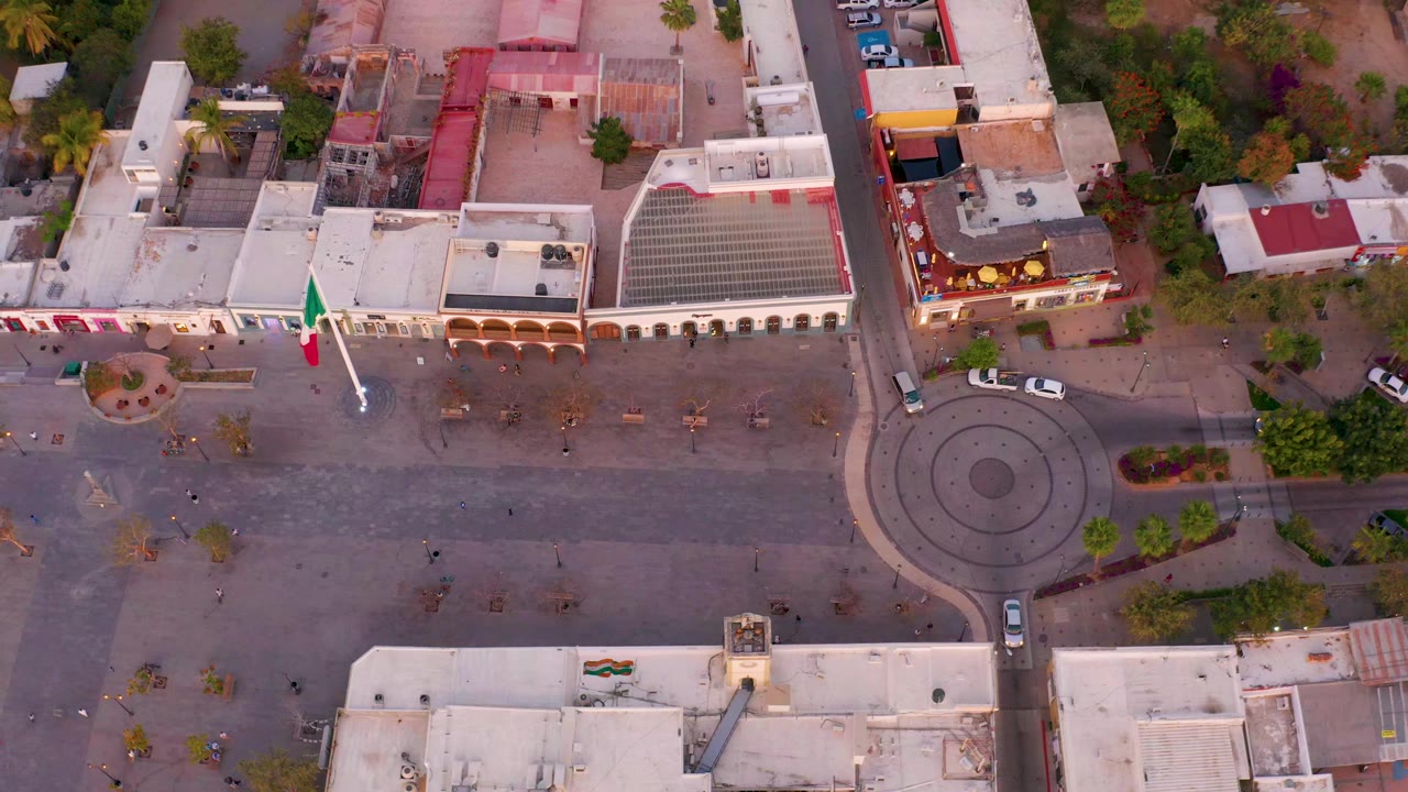 High aerial frame over San Jose Del Cabo, Mexico, showing a paved town square, low buildings, a flagpole, trees, and a roundabout with no visible people.