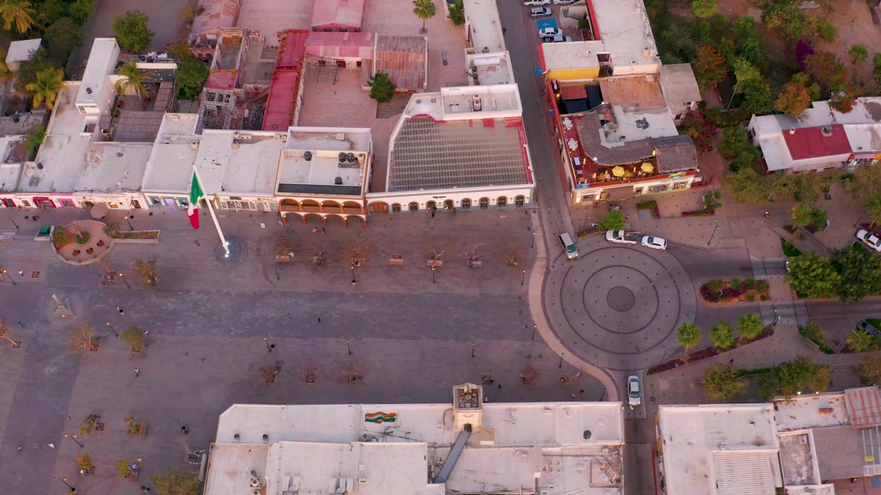 Aerial view of San Jose Del Cabo, Mexico, with the roundabout centered lower right, low commercial buildings across the top, trees, parked cars, and no visible people.