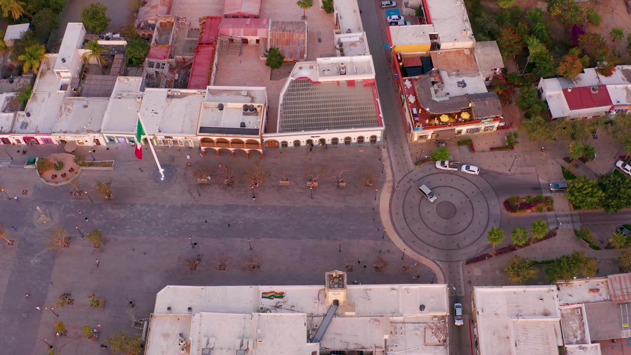 Aerial frame in San Jose Del Cabo, Mexico, with a car visible in the roundabout, a broad paved plaza, arched building frontage, trees, rooftops, and no visible people.