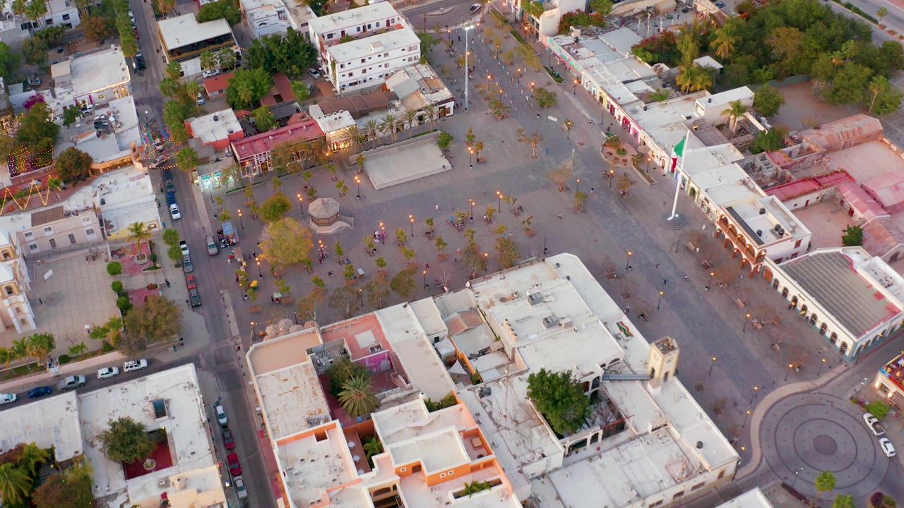 Overhead view of a San Jose del Cabo plaza with pale rooftops, street edges, scattered trees, parked cars, and small visible people near the square.