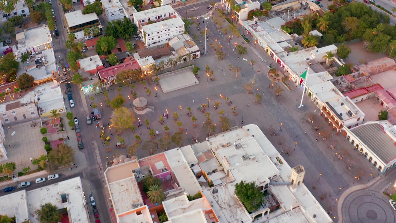 San Jose del Cabo aerial view with the Mexican flag near the right side, an arcaded white building along the plaza, parked cars on the street, trees, rooftops, and small visible people.