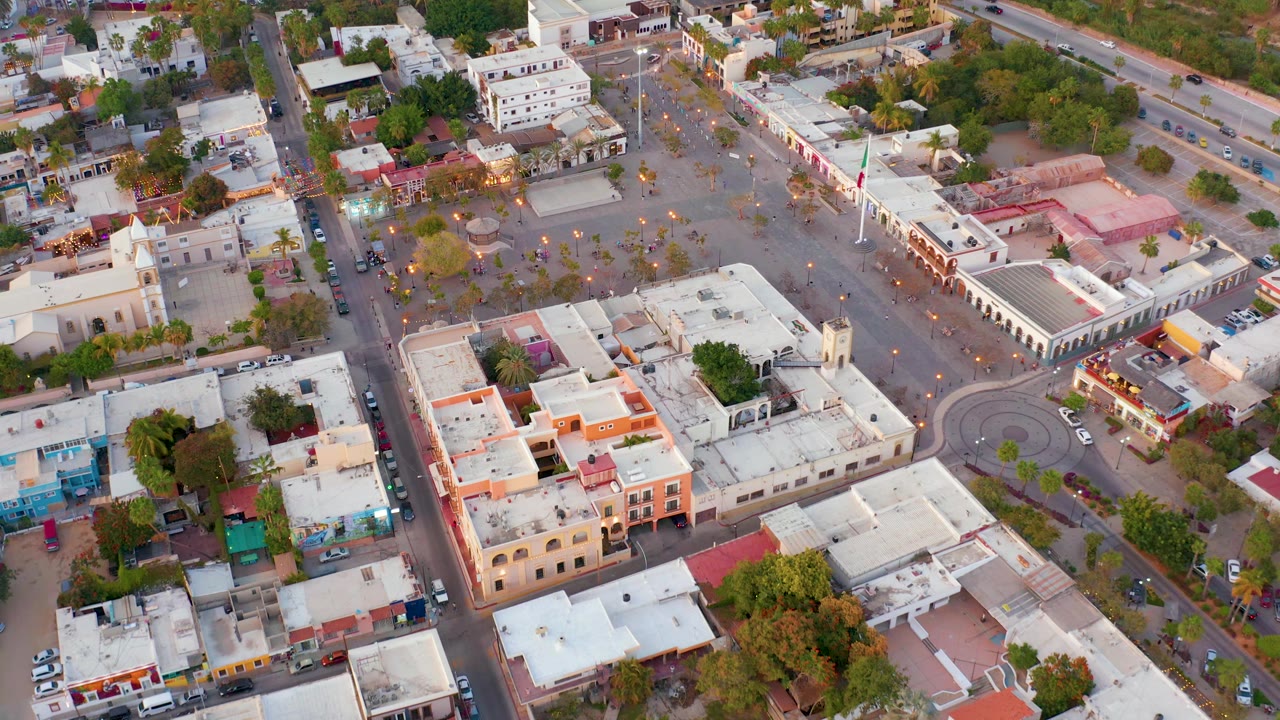 Descending drone view over San Jose del Cabo with the city square centered higher in frame, white rooftops in the foreground, street traffic at the edges, and no visible people.