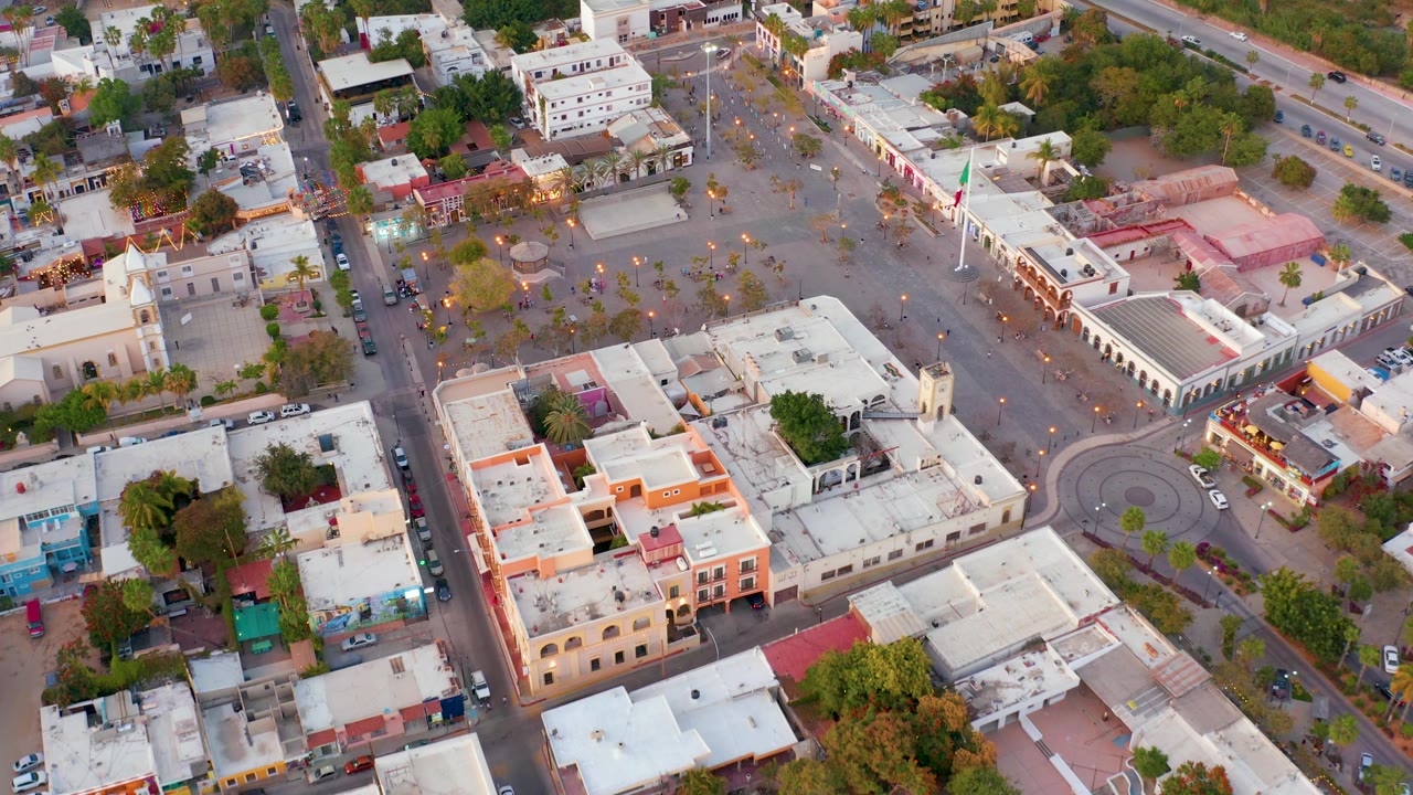 Aerial frame over San Jose del Cabo showing the plaza, flagpole, circular street feature, white-roofed buildings, scattered trees, cars, and no visible people.
