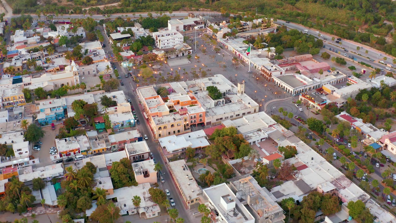 Aerial view in San Jose del Cabo, Mexico with the paved square higher in frame, compact rooftops below, roadside trees, parked cars, and no visible people.