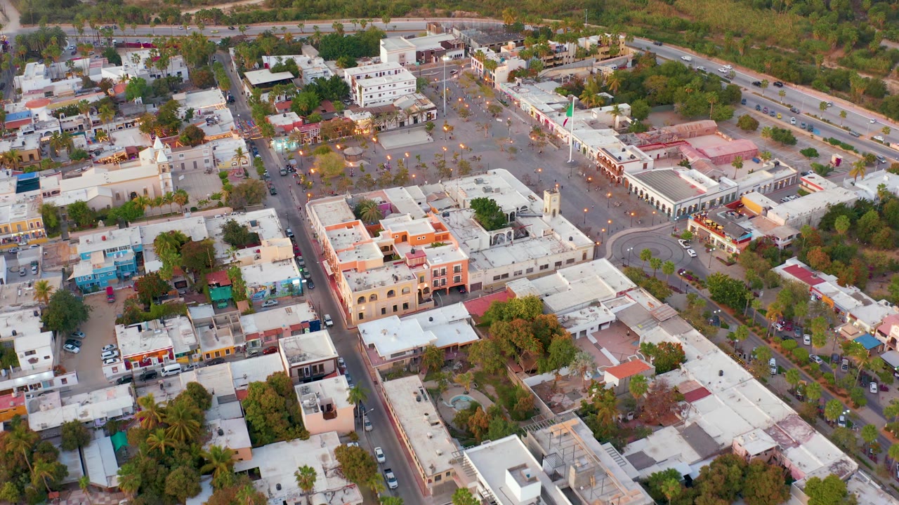 Drone-style aerial frame over San Jose del Cabo, Mexico with a roundabout near the right side, the paved town area in the upper center, rows of rooftops, cars, trees, and no visible people.