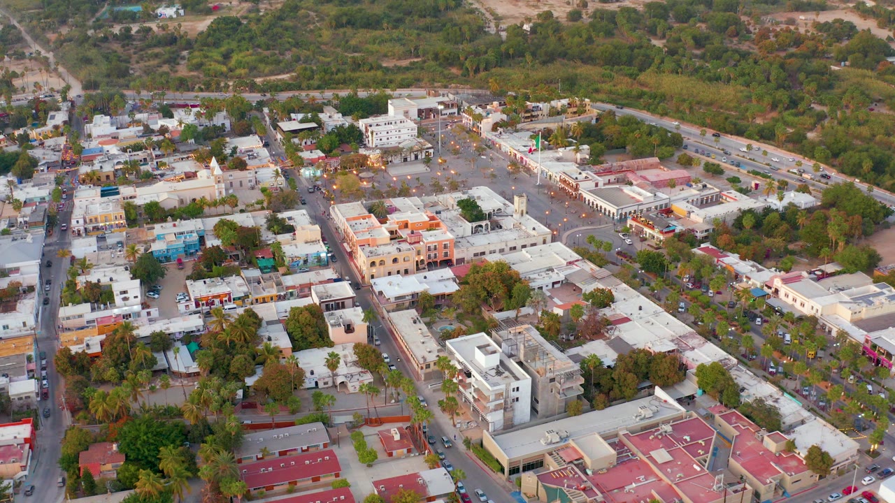 Oblique aerial frame over San Jose del Cabo showing low-rise town buildings, red and white rooftops, tree-lined streets, parked vehicles, and no visible people.