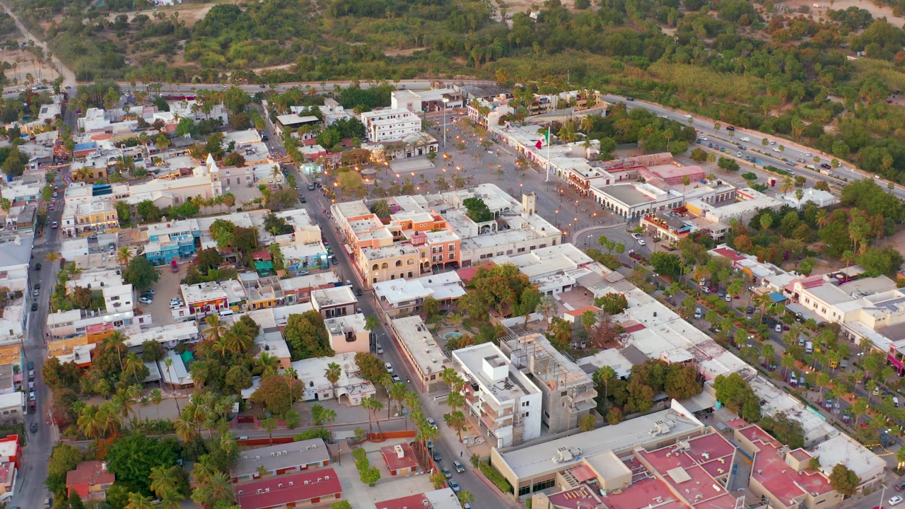 San Jose del Cabo appears from an elevated drone angle with pale buildings, red roof sections, street traffic, parked vehicles, dense trees, and no visible people.
