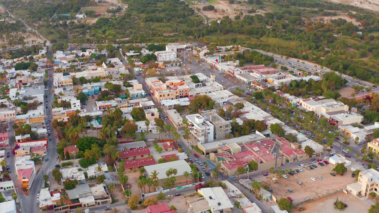 Opening aerial view over San Jose del Cabo town blocks with a vertical road at left, red-roofed buildings near the bottom, parking areas, tree cover, and no visible people.