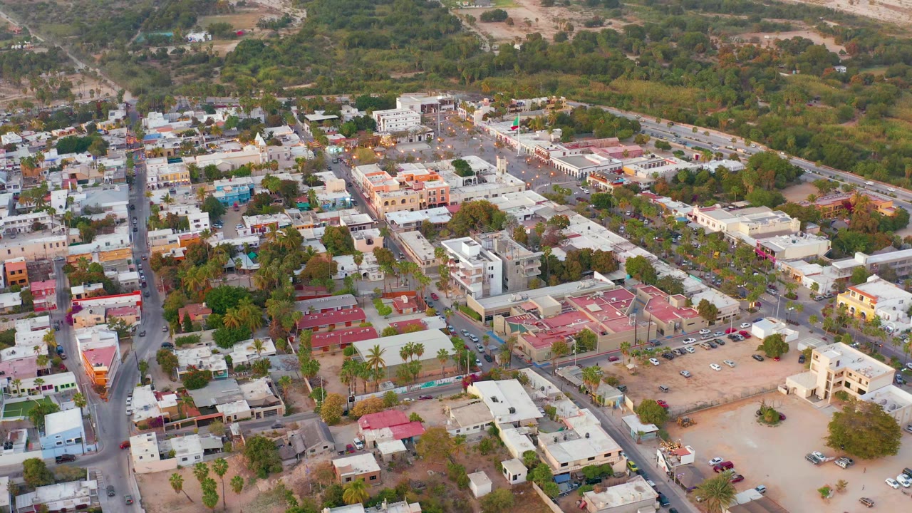 Aerial frame of San Jose del Cabo showing a diagonal road, clustered homes and commercial buildings, red roof sections, scattered vehicles, tree cover, and no visible people.