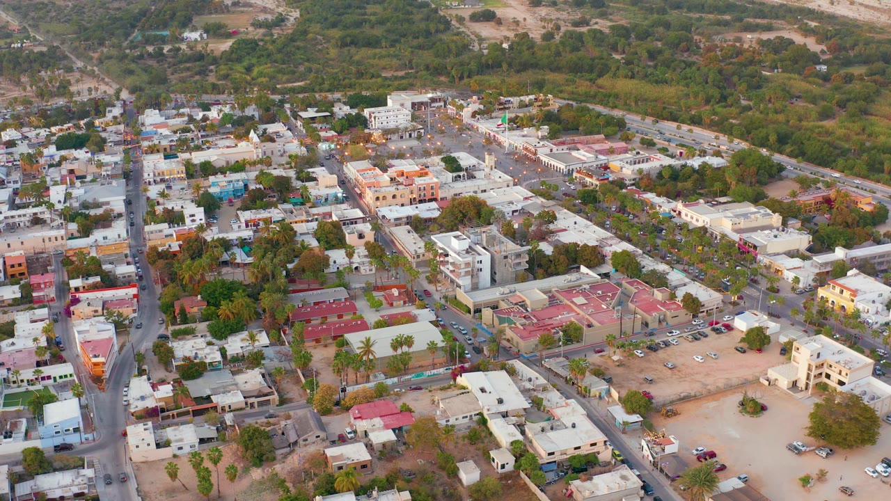 Oblique drone view across San Jose del Cabo streets, with a main road running through the left side, pale and red rooftops, parked vehicles, open dirt lots, and no visible people.
