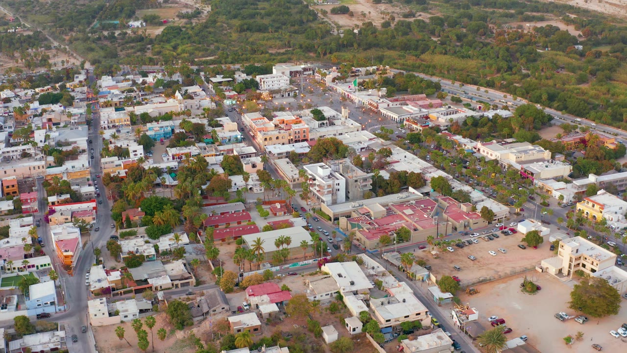 Aerial town scene with traffic, trees, pale buildings, red roofs, and hillside background.