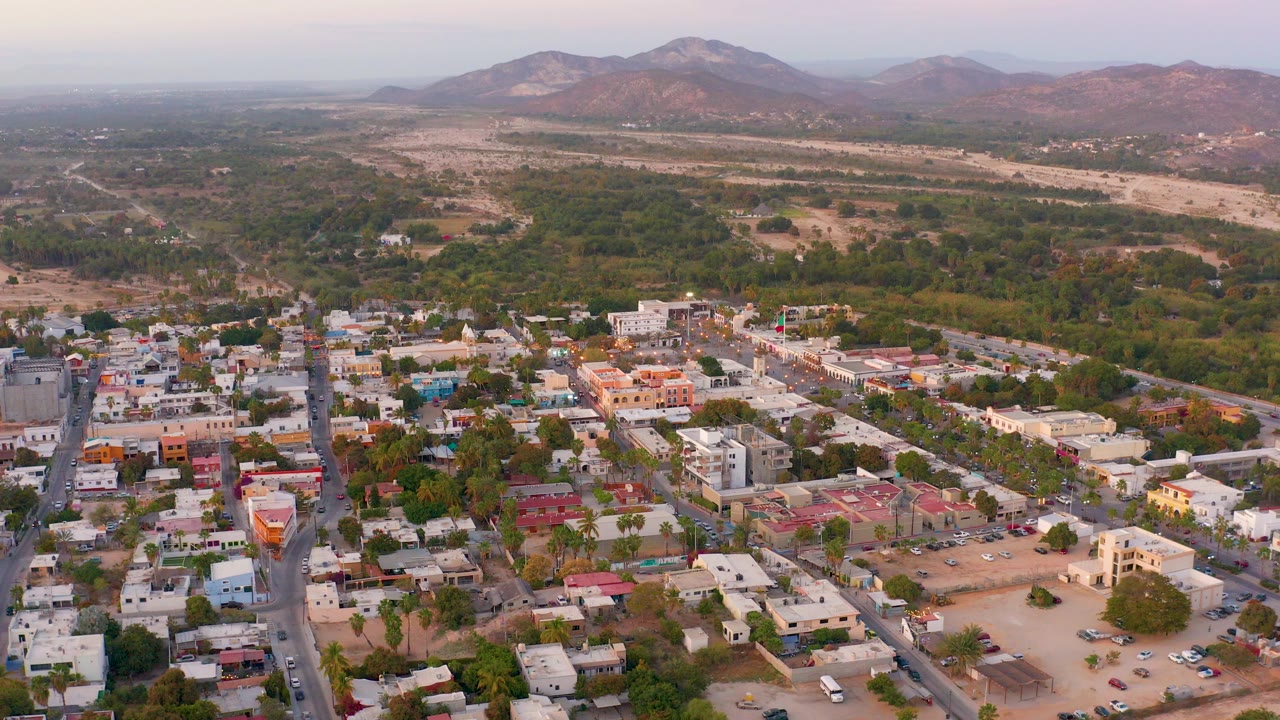 Opening aerial frame over San Jose del Cabo low-rise homes, red-roofed buildings, gridded streets, trees, dry open land, and distant mountains with no visible people.